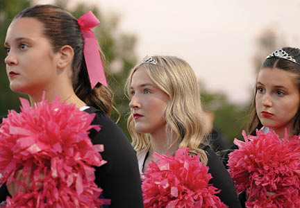 Three people wearing tiaras and holding pink pom-poms appear focused and are dressed in black outfits with one person having a pink ribbon in their hair. Description generated by AI