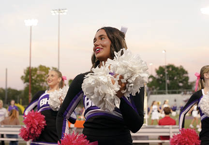 People in cheerleading uniforms hold pom-poms and perform outdoors near bleachers during dusk. Description generated by AI