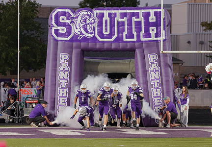 People in purple football uniforms are running out of an inflatable tunnel labeled “South Panthers" onto a football field with smoke effects. Description generated by AI