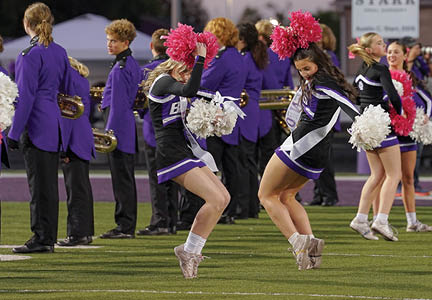 Two people in cheerleading uniforms with pom-poms are energetically performing on a field with a marching band in purple uniforms standing in the background. Description generated by AI