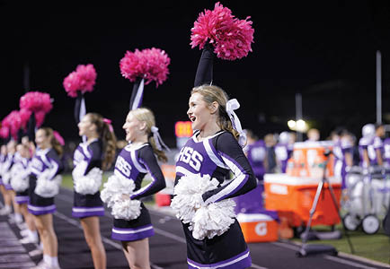 A group of cheerleaders in matching purple and white uniforms are performing with pom-poms on a sports field at night. Description generated by AI