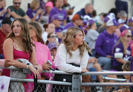 People wearing casual clothing are gathered in a stadium seating area, with some leaning on a railing and others sitting in the background. Description generated by AI