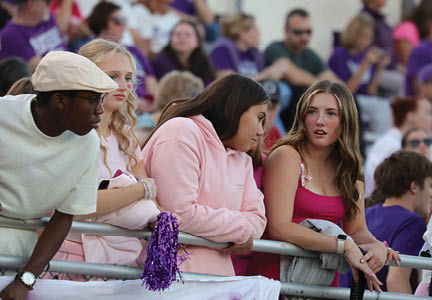 Four people wearing casual clothing are leaning on a railing at an outdoor event with a crowd in the background. Description generated by AI