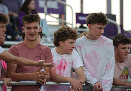 A group of young people wearing casual clothes, some with pink stains, are standing and leaning on a railing while engaging in conversation at an outdoor event. Description generated by AI