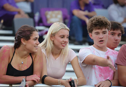 Four people are standing close together at an outdoor event, with three of them engaged in conversation and one gesturing with an outstretched arm. Description generated by AI