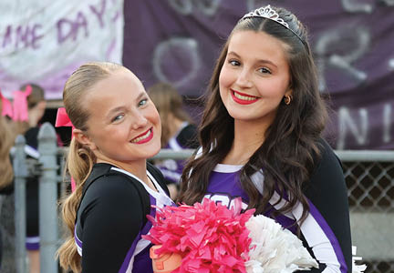 Two smiling adults with styled hair and red lipstick pose closely together in front of a crowd and a banner at an outdoor event. Description generated by AI