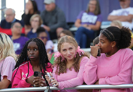 Three people wearing pink tops are standing behind a railing, with one holding a phone and another holding a snack, while a crowd is visible in the background. Description generated by AI