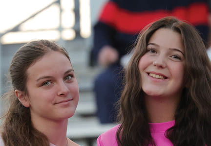 Two smiling people with long hair are in the foreground while a seated person wearing a dark sweater with a red stripe is in the background. Description generated by AI