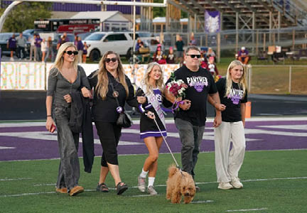 Five people walking arm in arm on a sports field with one person holding a bouquet and a small dog on a leash walking in front of them. Description generated by AI