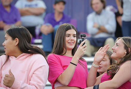A group of people wearing casual clothing are interacting and smiling in an outdoor setting with some seated in the background. Description generated by AI