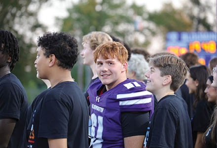 A group of people stand outdoors with one person in a purple sports jersey smiling at the camera while others in black shirts look in different directions. Description generated by AI