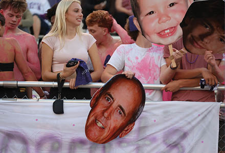 A large cutout of a smiling adult's face is held above a white banner with partially visible purple text behind a metal fence. Description generated by AI