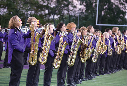 A line of people dressed in purple jackets and black pants are standing on a field playing saxophones. Description generated by AI