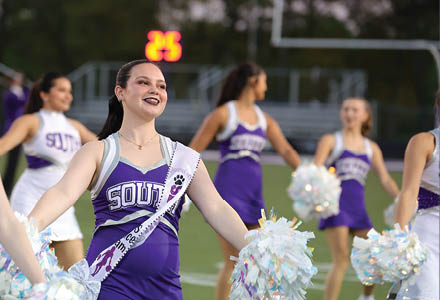 A group of adults in purple and white cheerleading uniforms are performing with pom-poms on a sports field. Description generated by AI