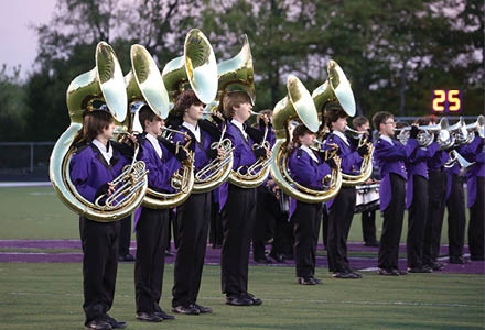 People in purple uniforms are standing in a line on a field playing brass instruments during a band performance. Description generated by AI