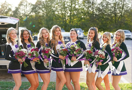 A group of eight adults dressed in matching cheerleading uniforms stand outdoors holding bouquets of flowers and smiling. Description generated by AI