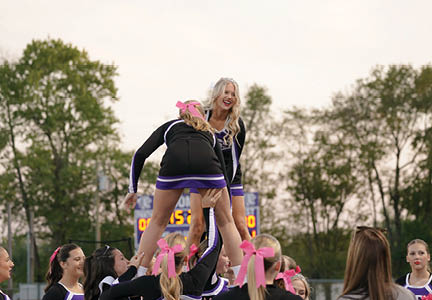 A group of people in cheerleading uniforms with pink bows are performing a stunt outdoors with one person lifted above the others smiling. Description generated by AI