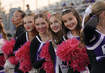 Two people wearing cheerleading uniforms hold pink pom-poms, with one person having long curly hair. Description generated by AI