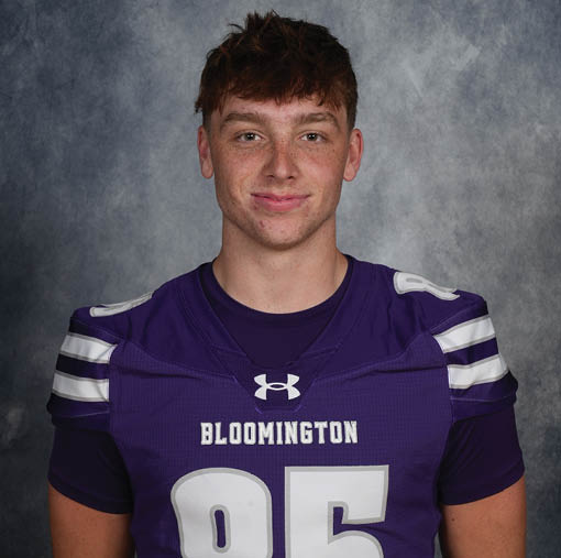 A person with short, tousled hair is wearing a purple Bloomington football jersey with the number 85 and is smiling slightly against a gray mottled background. Description generated by AI