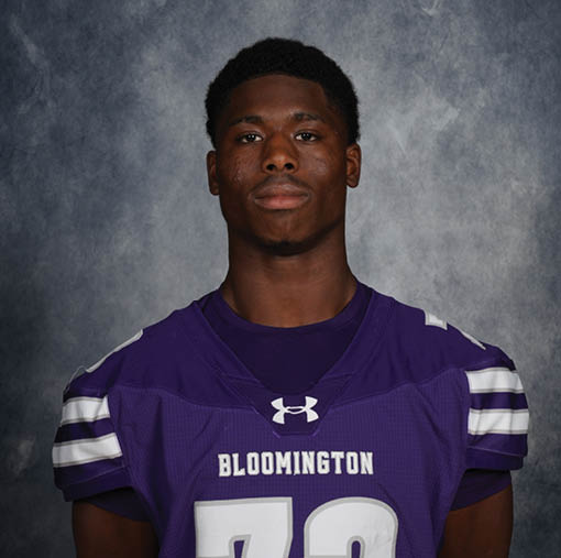 A person wearing a purple Bloomington sports jersey with the number 72 stands against a gray textured background. Description generated by AI
