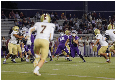 A group of people in football uniforms are engaged in a nighttime game on a field with spectators in the background. Description generated by AI
