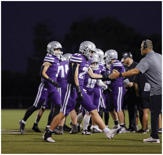 People in purple sports uniforms and helmets are walking on a field while a person in casual clothing gestures toward them. Description generated by AI