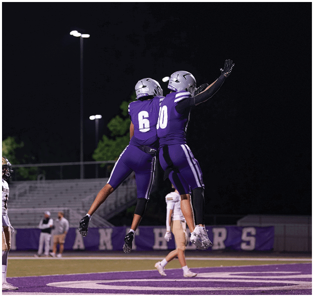 Two people in football uniforms are on a field at night with empty bleachers and a few spectators in the background. Description generated by AI