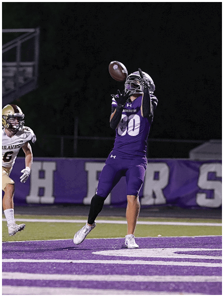 A person wearing a football uniform with the number 6 and a helmet is running on a field with empty bleachers and trees in the background. Description generated by AI