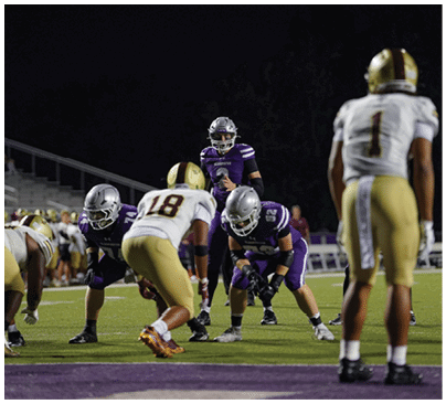 People in football uniforms are positioned on a field at night, preparing for a play during a game. Description generated by AI