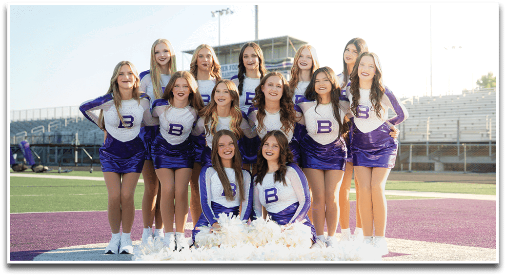 A group of people wearing matching purple and white cheerleading uniforms pose together on a sports field with smiles. Description generated by AI