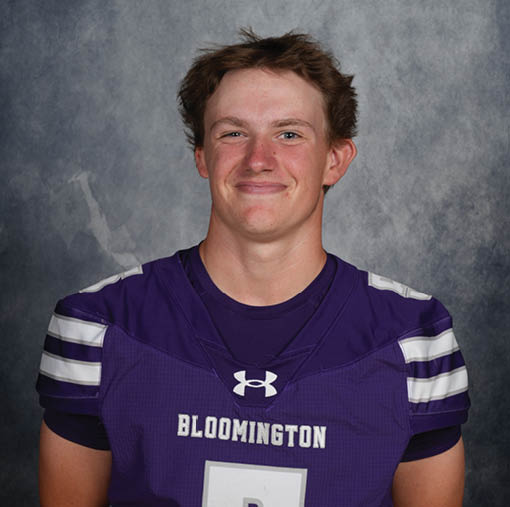A person with short, tousled hair is smiling and wearing a purple sports jersey with the word “BLOOMINGTON" on the front against a gray mottled background. Description generated by AI