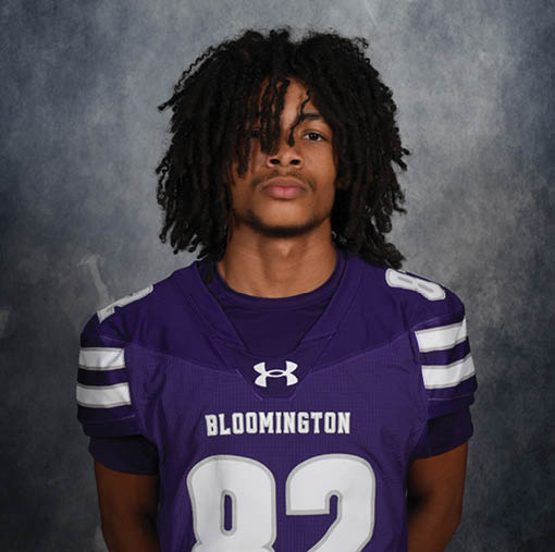Person with long curly hair wearing a purple Bloomington sports jersey with the number 82 stands against a gray textured background. Description generated by AI