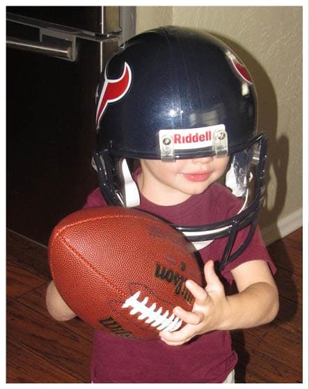 A child wearing a football helmet and a maroon shirt is holding a football indoors on a wooden floor. Description generated by AI