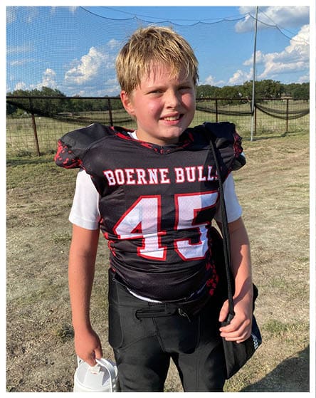 A child wearing a football uniform with the number 45 and the words “Boerne Bulls" is standing outdoors holding a water jug and a black bag. Description generated by AI