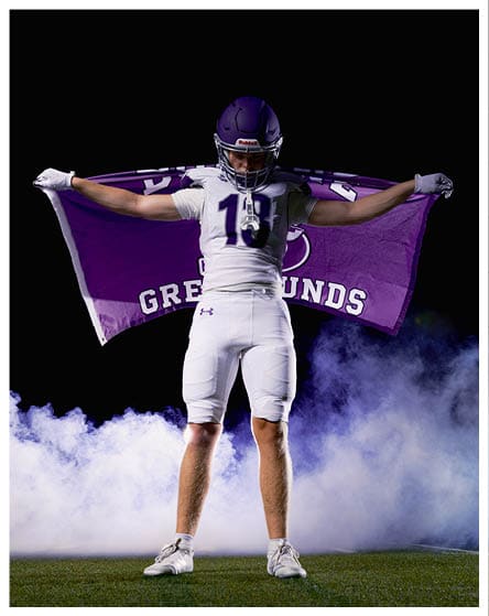 A person in a white football uniform stands on a field holding a purple flag with the word “GREYHOUNDS" spread out behind them against a smoky dark background. Description generated by AI