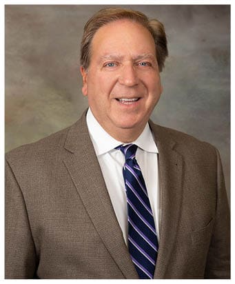A person wearing a brown textured suit jacket, white dress shirt, and a blue and purple striped tie is smiling. Description generated by AI