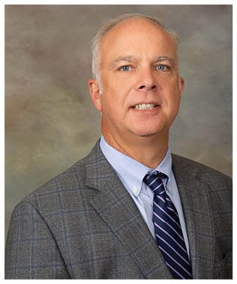 A person wearing a gray plaid suit jacket, a light blue button-down shirt, and a navy blue tie with diagonal light purple stripes. Description generated by AI