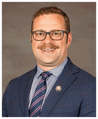 A person wearing a striped shirt, a dark suit jacket with a subtle check pattern, a striped tie, and a gold lapel pin is smiling. Description generated by AI