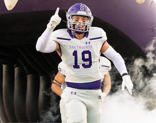 A person in a white and purple football uniform with the number 19 is running out of a tunnel through smoke while raising one finger. Description generated by AI