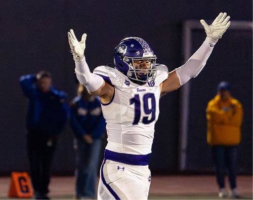 A person in a white and purple football uniform with the number 19 is raising both arms on a football field with blurred people and an orange marker in the background. Description generated by AI