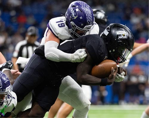Two people in football uniforms are engaged in a tackle on a field, with one holding the ball tightly while the other wraps their arms around them. Description generated by AI