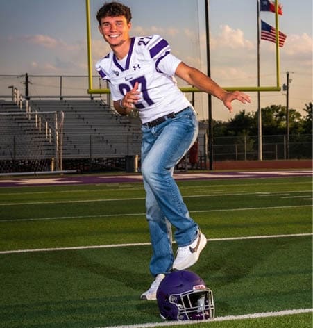 A person wearing a white sports jersey with the number 27 and blue jeans is in a dynamic pose on a sports field with bleachers in the background. Description generated by AI