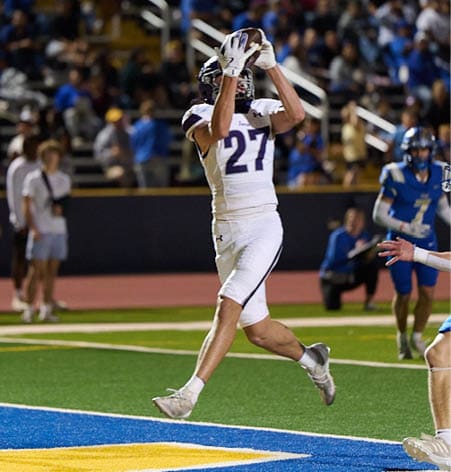 A person wearing a white sports uniform with the number 27 is jumping to catch a football on a field while other players in blue uniforms watch nearby. Description generated by AI