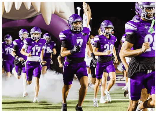 Three people in purple football uniforms and helmets are on a field at night, with one person raising an arm in the background. Description generated by AI