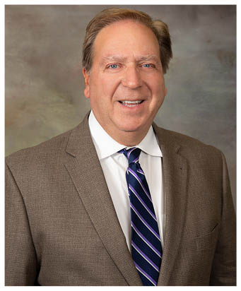 A person with short hair wearing a brown suit jacket, white shirt, and striped tie is smiling against a neutral background. Description generated by AI