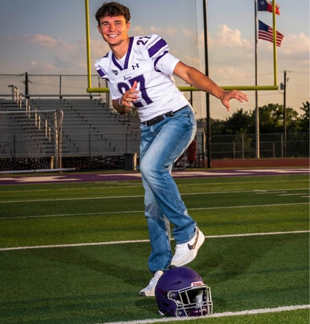 A person wearing a white sports jersey with the number 27 and blue jeans is in a dynamic pose on a sports field with bleachers in the background. Description generated by AI