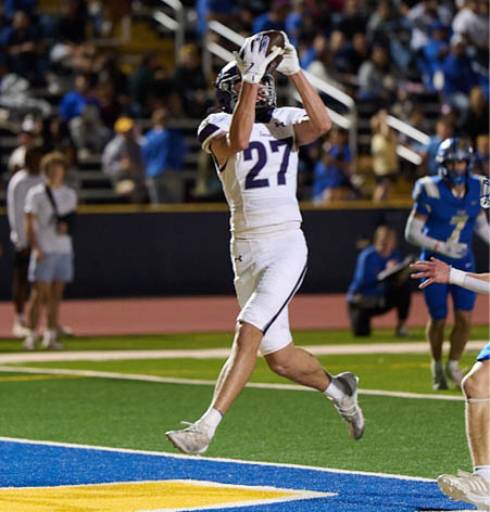 A person wearing a white sports uniform with the number 27 is jumping to catch a football on a field while other players in blue uniforms watch nearby. Description generated by AI