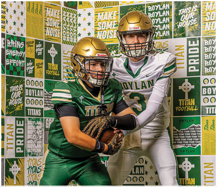 Two people wearing football helmets and uniforms pose in front of a wall covered with green and yellow sports-themed posters and slogans. Description generated by AI