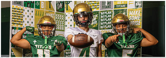 Three people wearing football uniforms stand side by side with two flexing their arms and the middle person holding a football. Description generated by AI