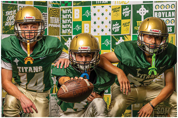 Three people wearing gold football helmets and green jerseys are positioned in front of a wall covered with green, white, and yellow sports-themed posters and signs. Description generated by AI